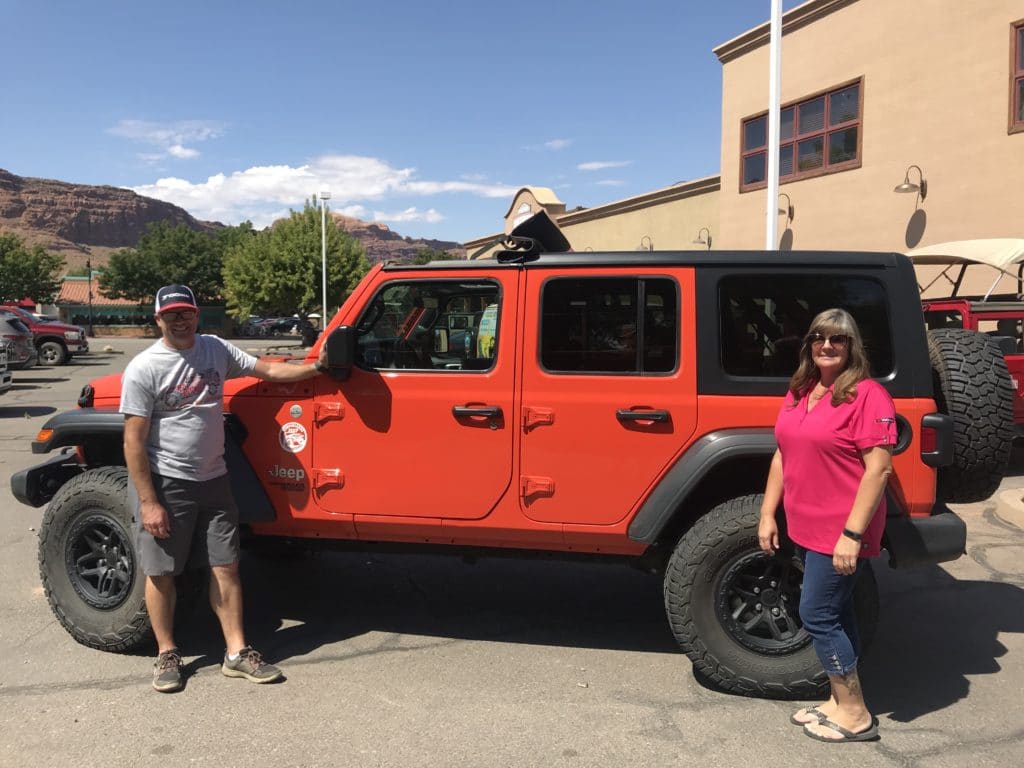 man and woman stand infront of red 4 door jeep one by the front wheel, the other by the back wheel